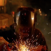 A welder produces sparks in a dark workshop, contributing to the output of a larger welding operation.